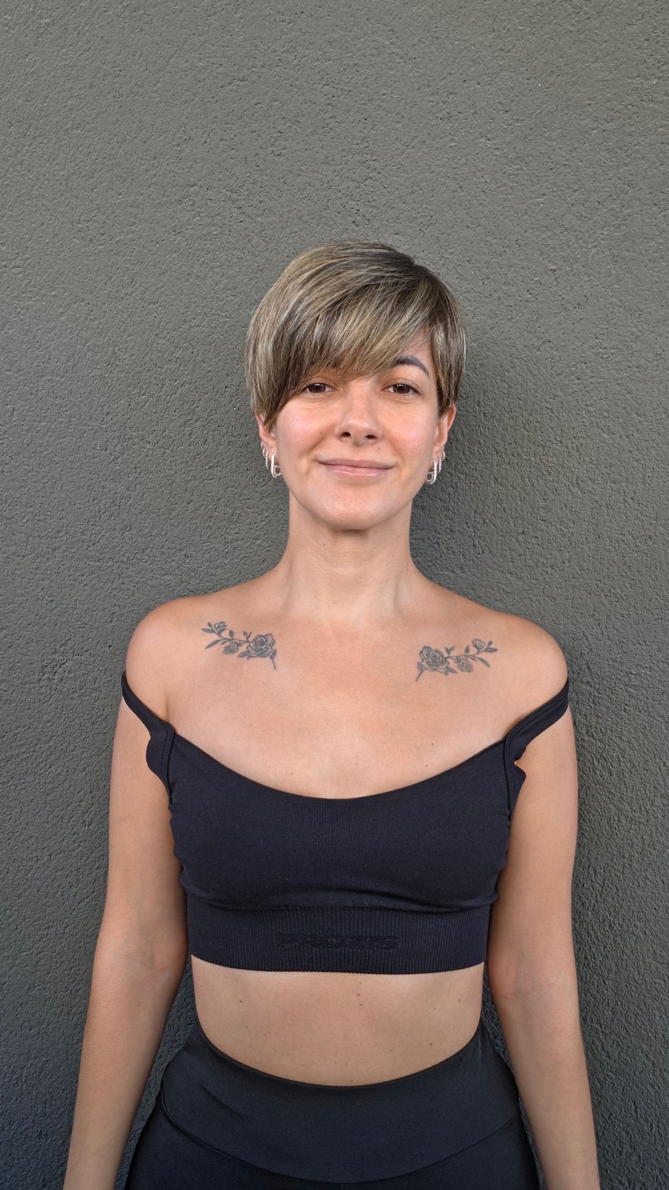 Retrato frontal de una mujer de pelo corto contra una pared gris, con top deportivo negro.