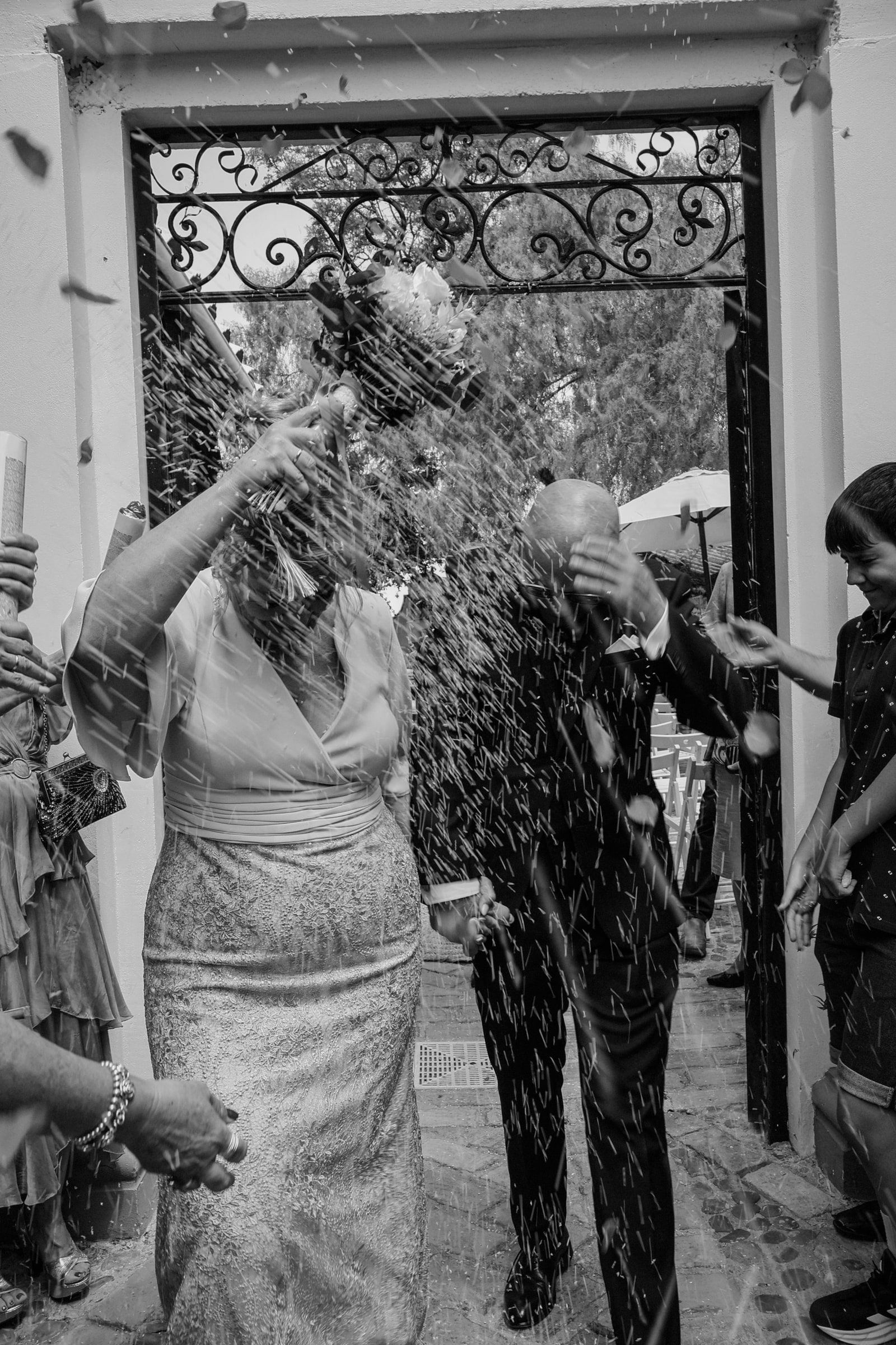 Blanco y negro: una pareja sale por una cancela de forja mientras los invitados les lanzan arroz.