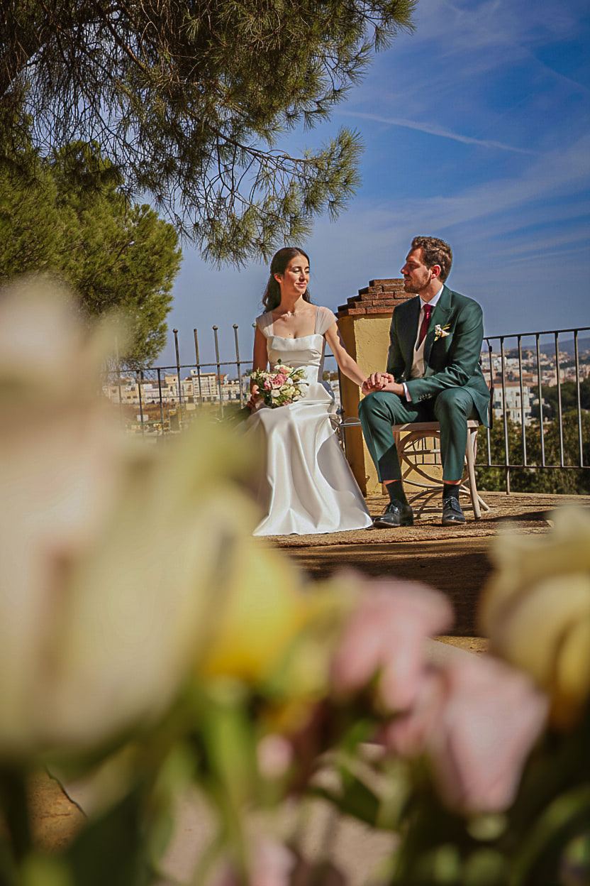 Los novios sentados en un mirador, vistos a través de unas flores desenfocadas en primer plano.