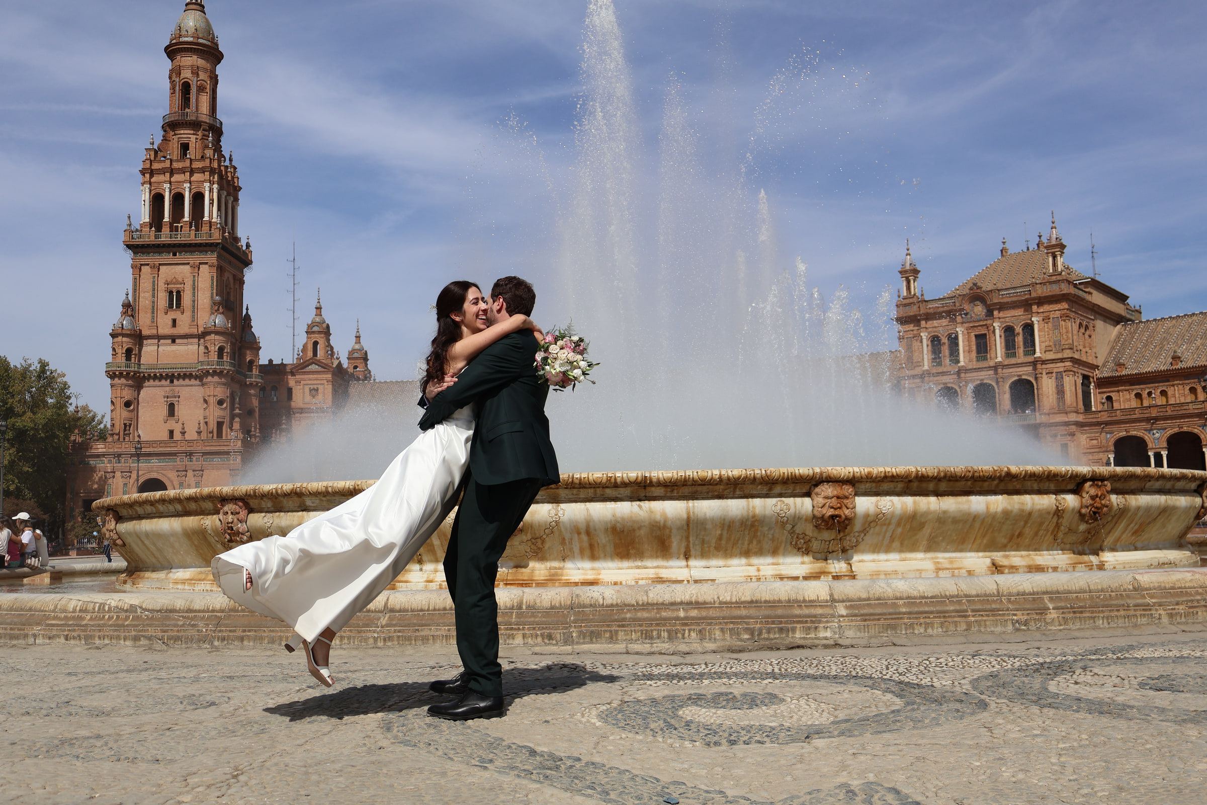 Un novio levanta en brazos a la novia delante de la fuente de la Plaza de España de Sevilla, con cielo azul.