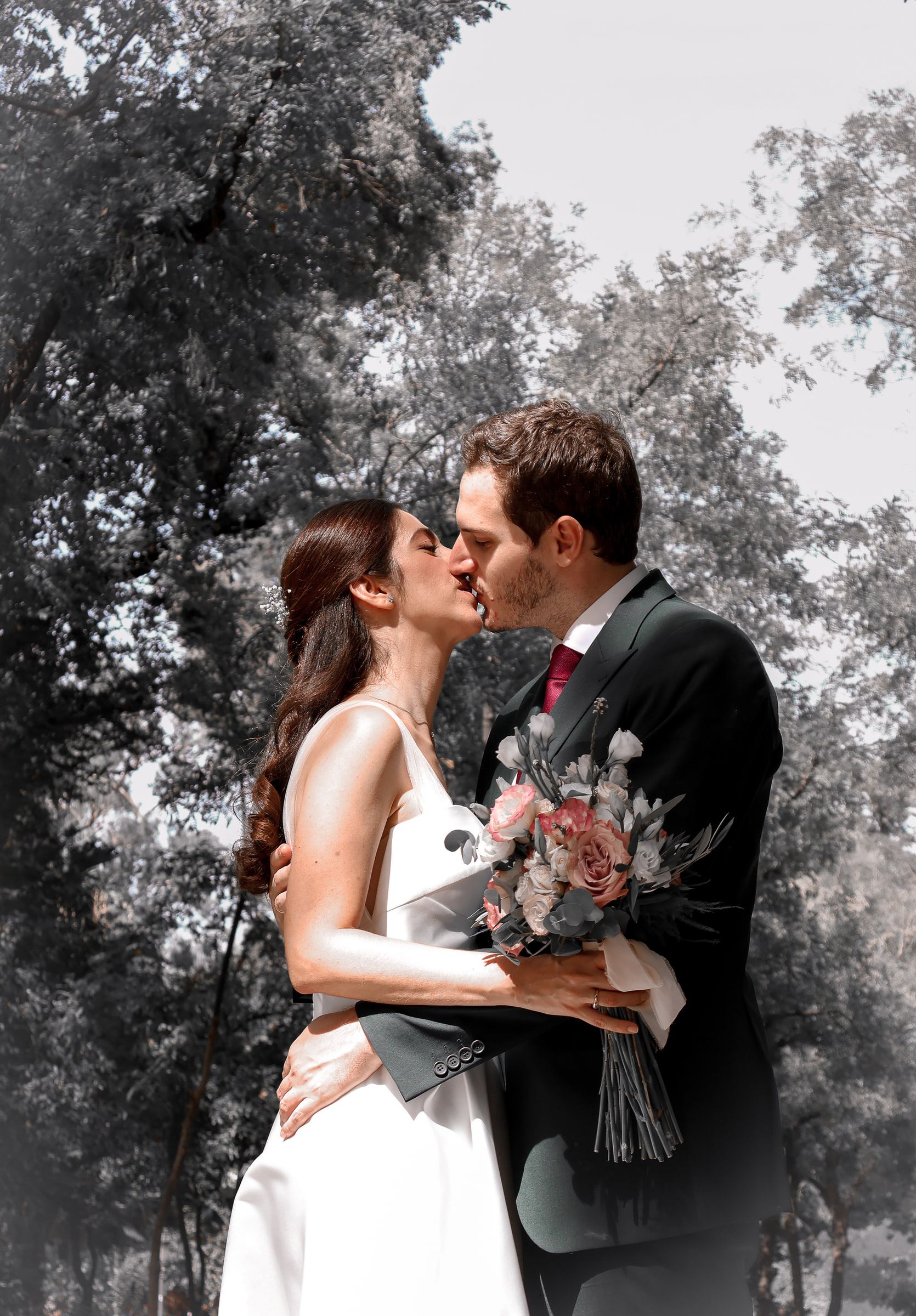 Los novios se besan bajo una arboleda, con el fondo virado a blanco y negro y ellos en color.