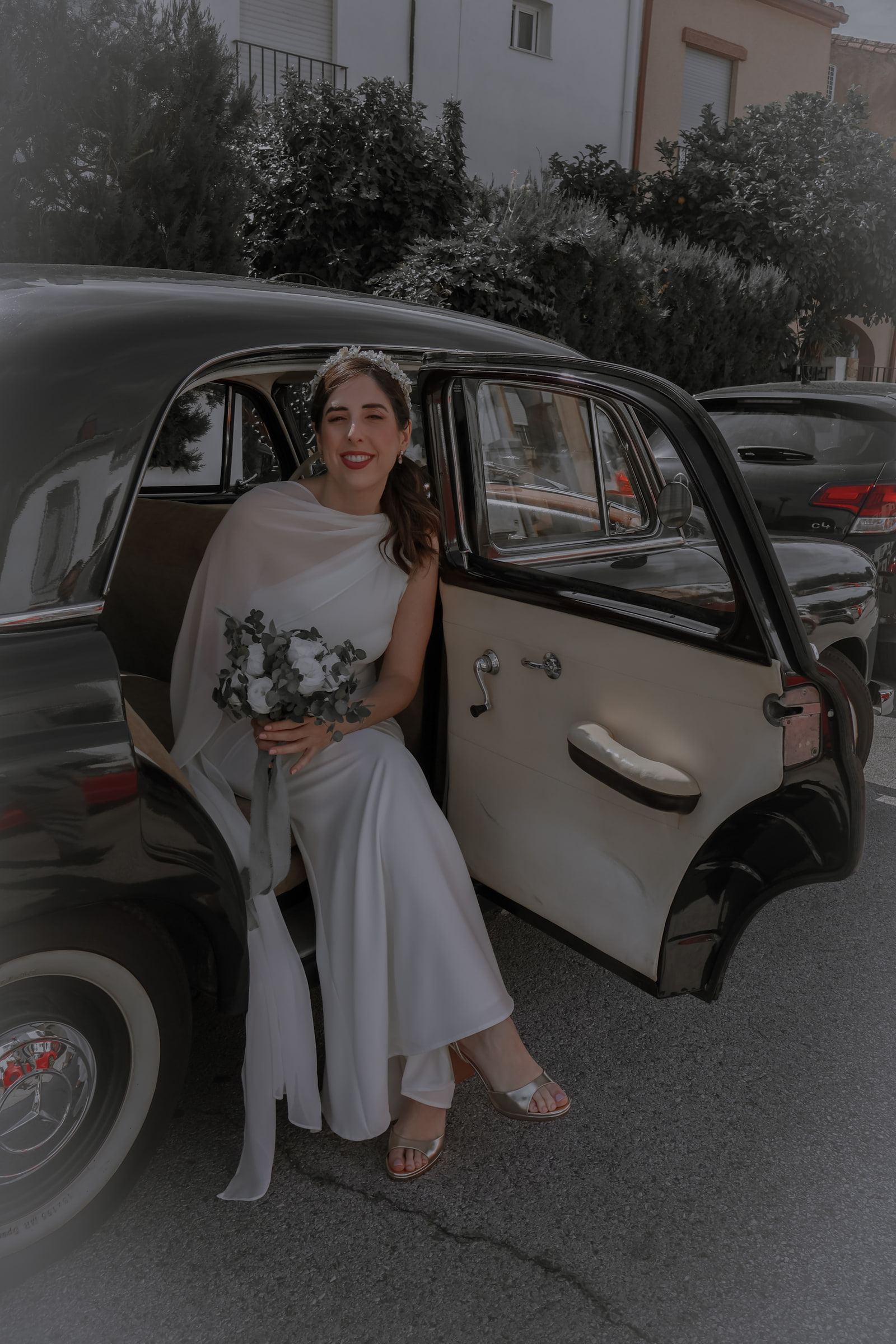La novia sentada en la puerta abierta de un coche clásico negro, con el ramo en el regazo.