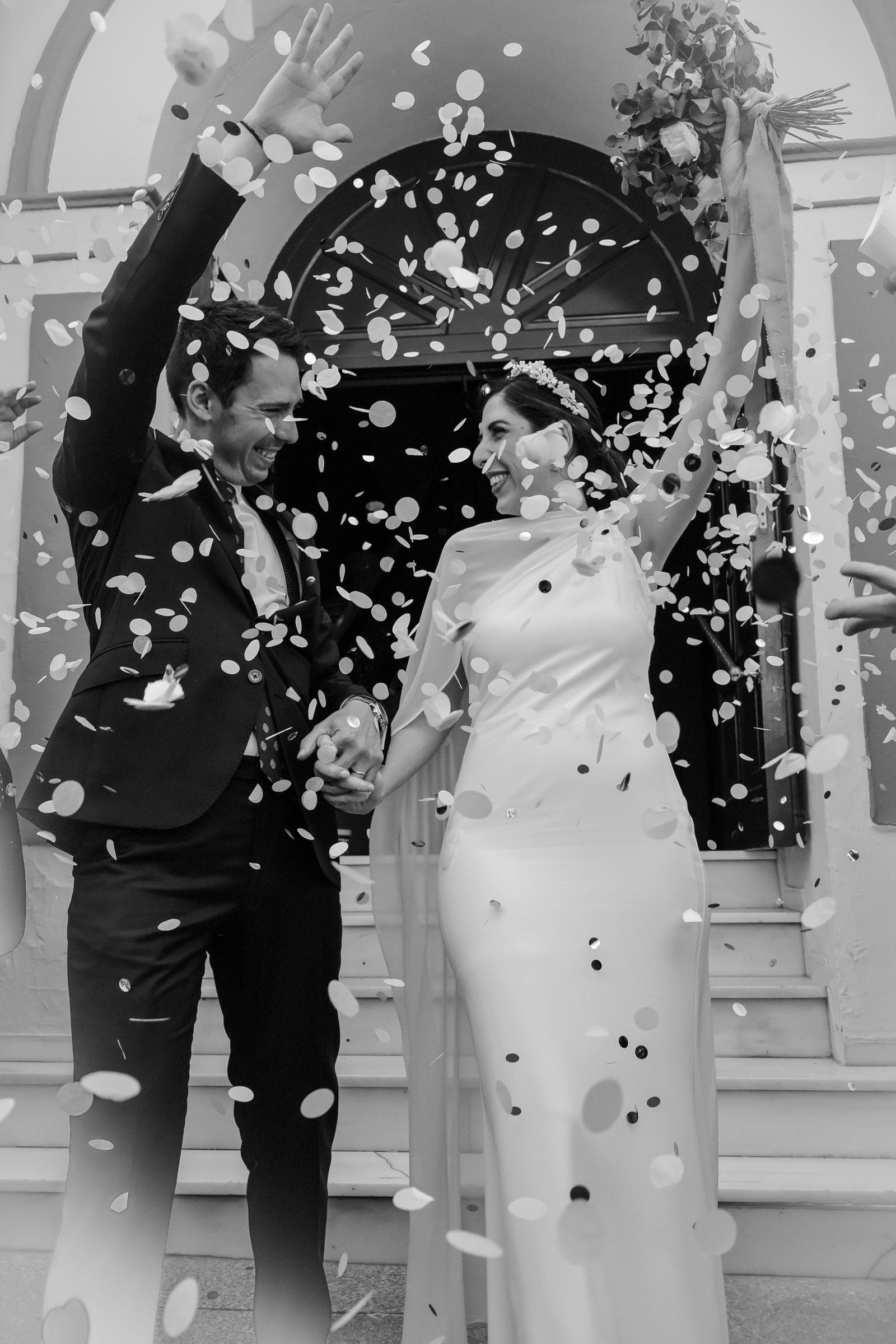 Blanco y negro: los novios salen de la ceremonia con los brazos en alto bajo una lluvia de confeti.