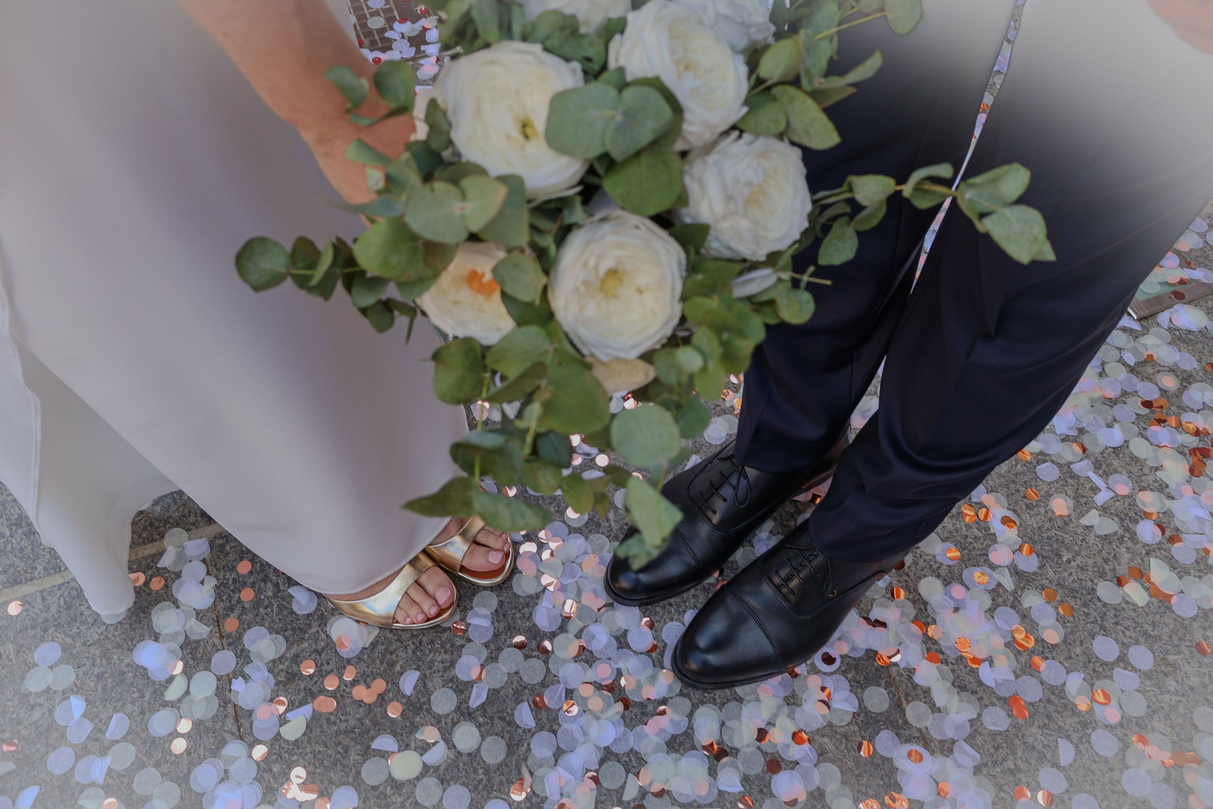 Detalle a ras de suelo: el ramo de rosas blancas y eucalipto entre los zapatos de los novios, sobre confeti de colores.