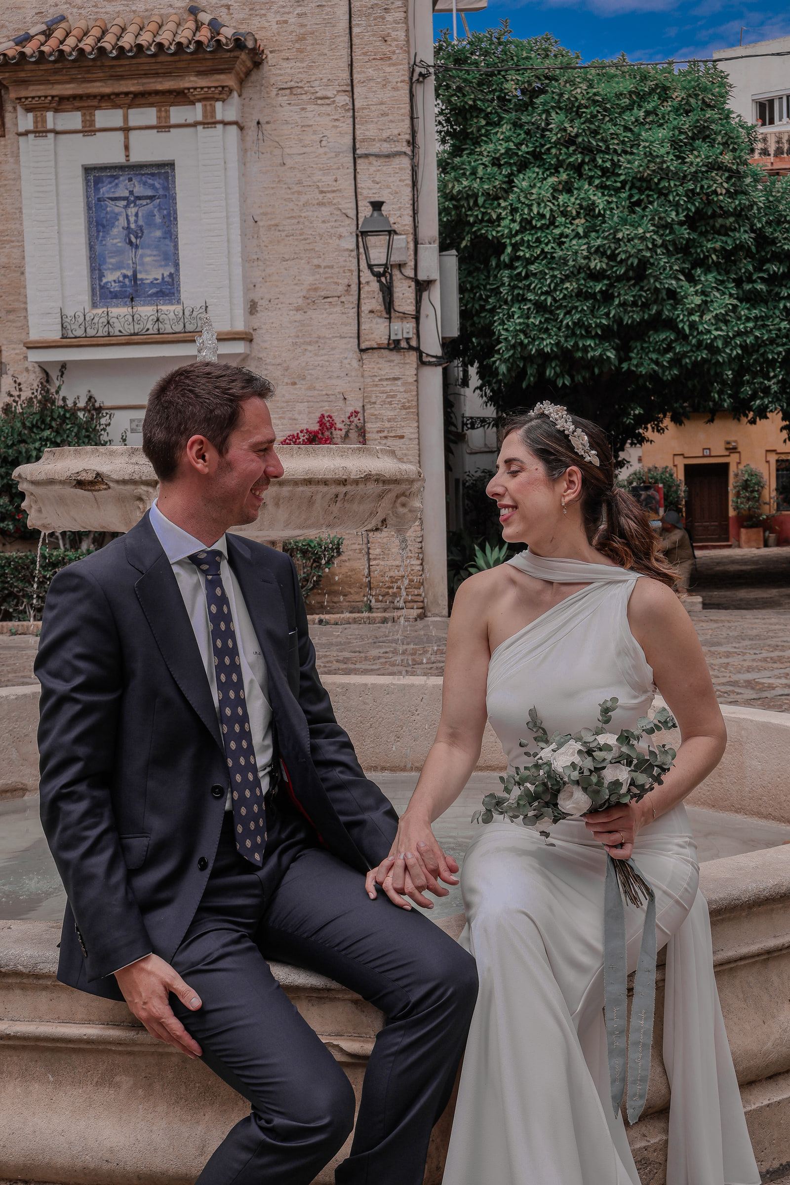 Los novios sentados en el borde de una fuente de una plaza sevillana, mirándose y riendo.