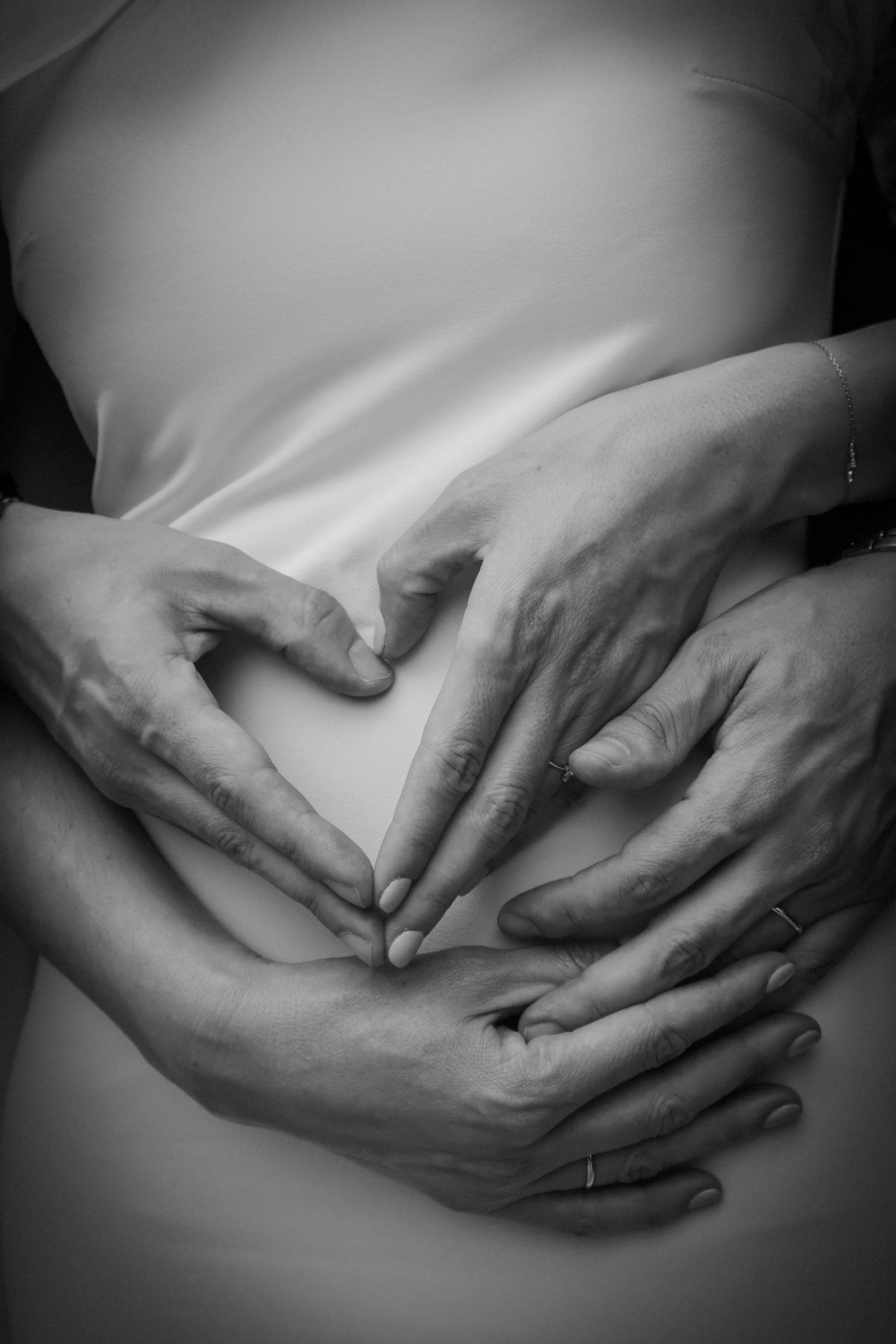 Blanco y negro: dos pares de manos forman un corazón sobre la barriga de una embarazada.