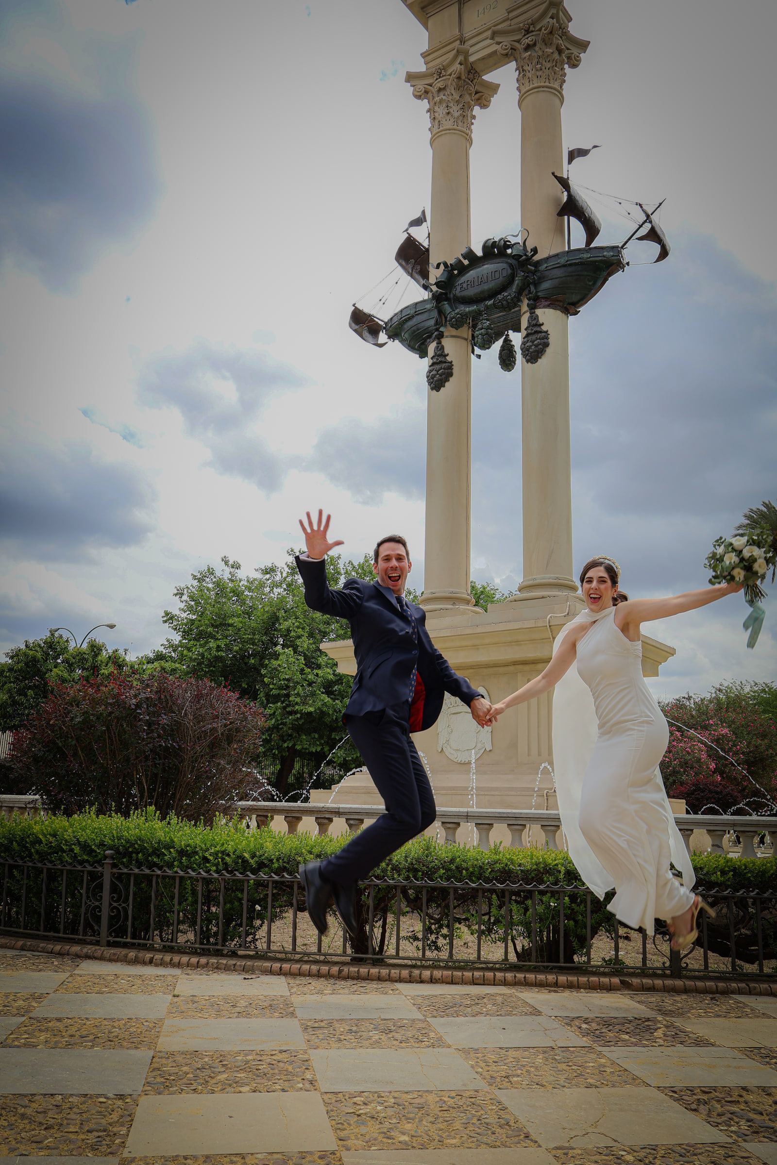 Los novios saltan de la mano delante de un monumento de columnas, con cielo nublado.