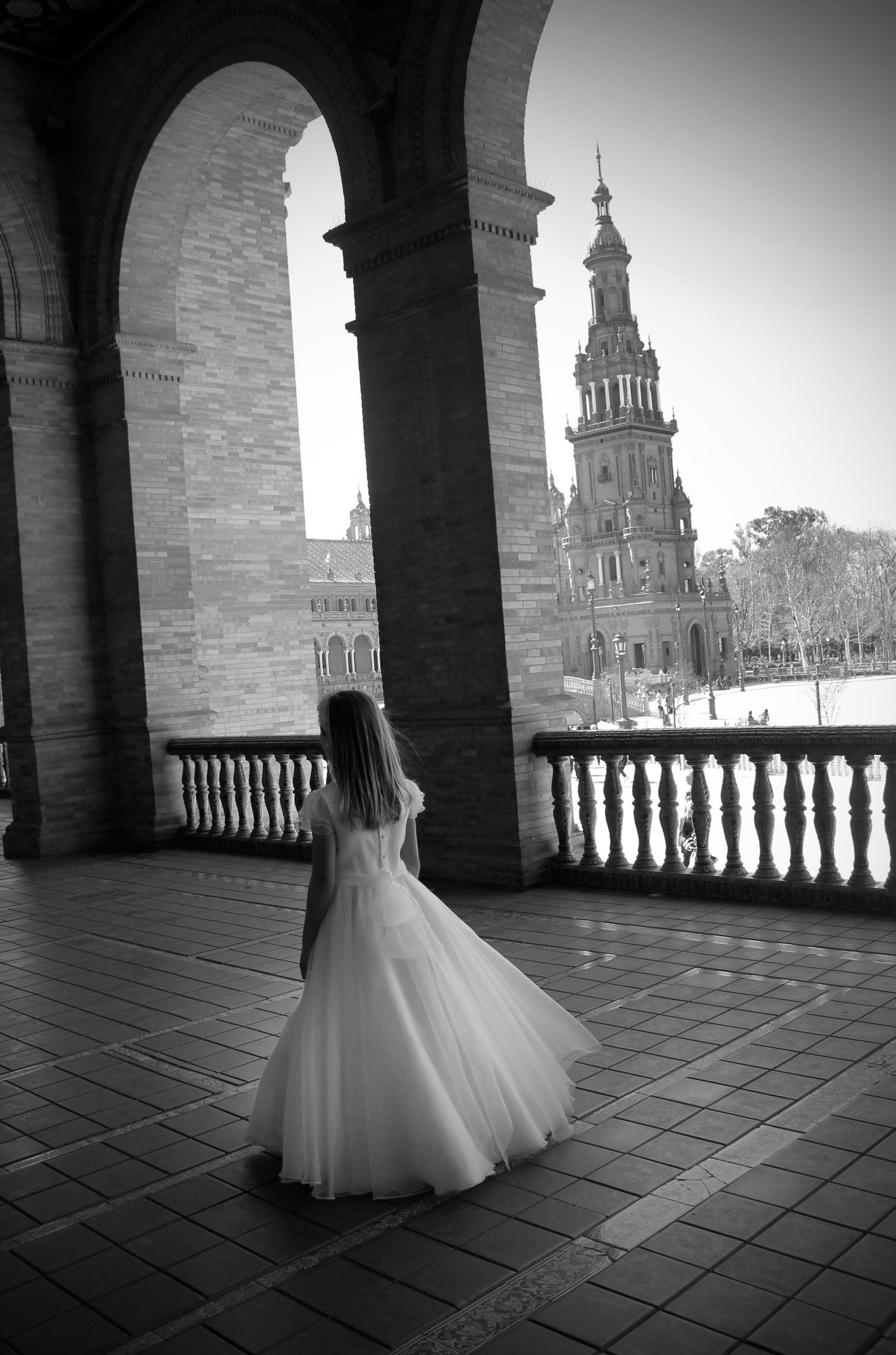 Blanco y negro: una niña con vestido de comunión, de espaldas, en la galería de la Plaza de España.