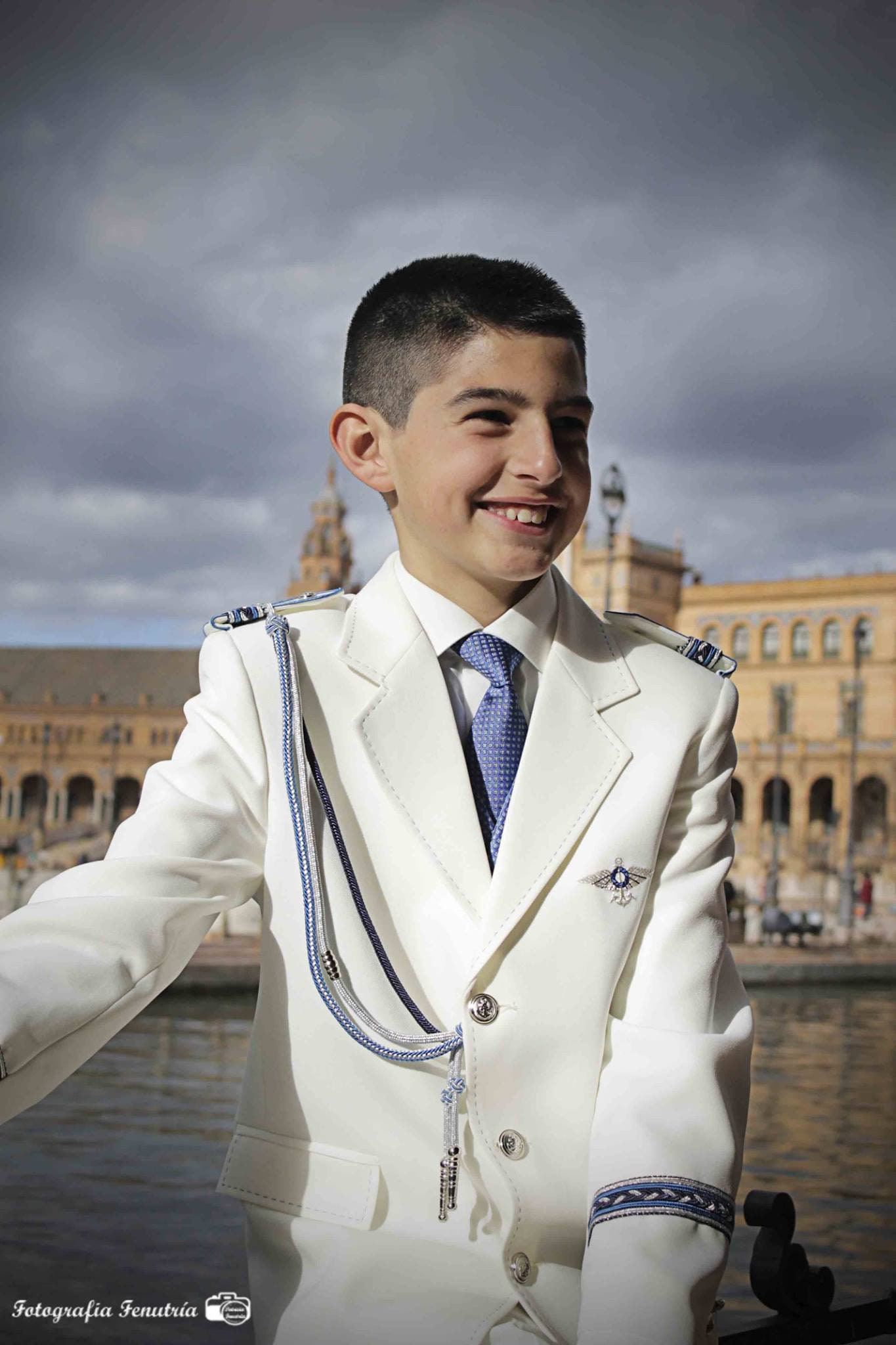 Retrato de un niño de comunión con traje marinero blanco, con la Plaza de España y un cielo de tormenta detrás.