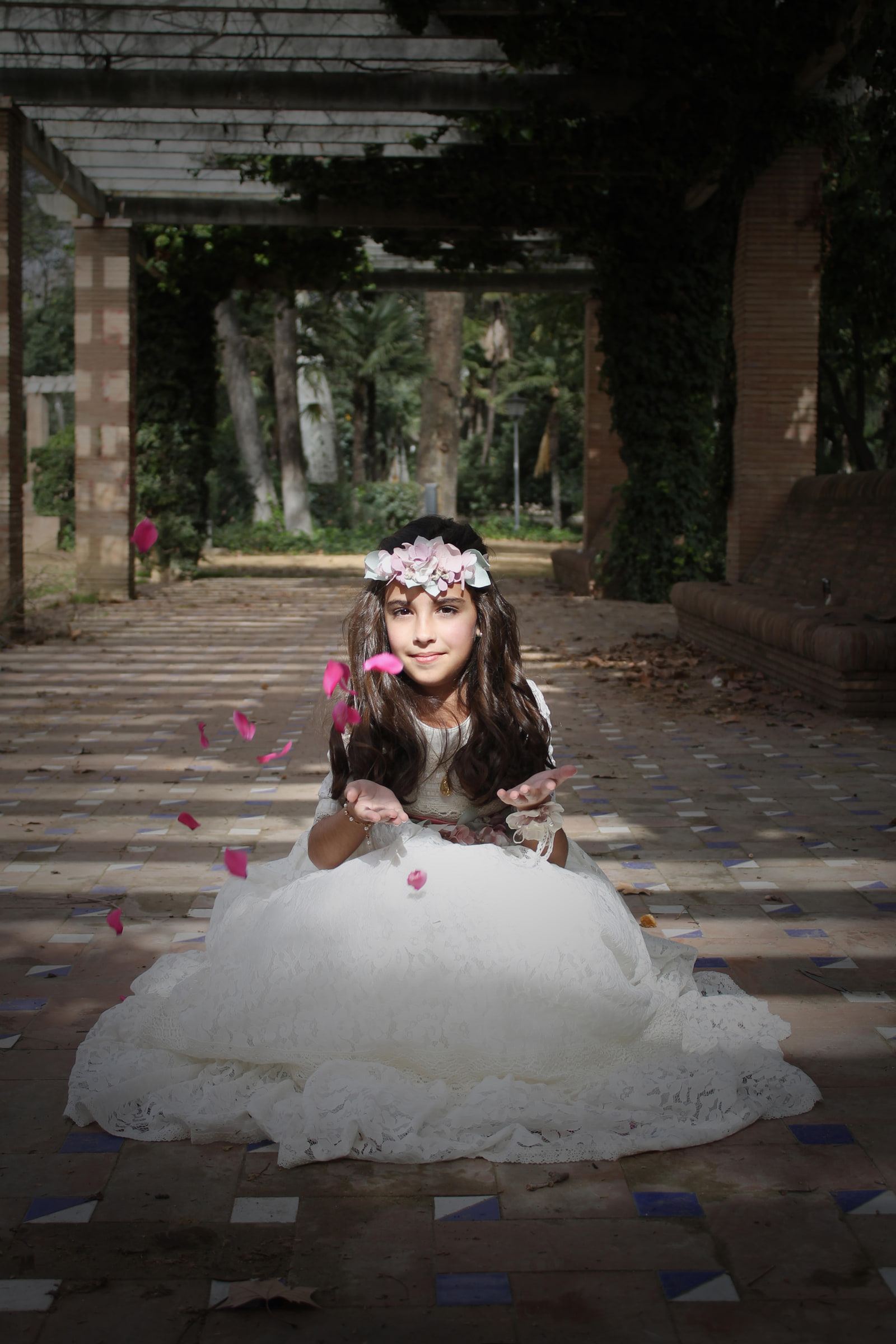 Una niña de comunión sentada en una pérgola ajardinada sopla pétalos rosas desde las manos.