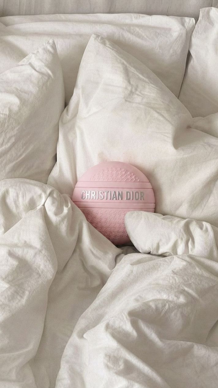 IA. Packshot de un bote de crema rosa sobre fondo gris y, generado con IA, el mismo bote entre sábanas y almohadas blancas.
