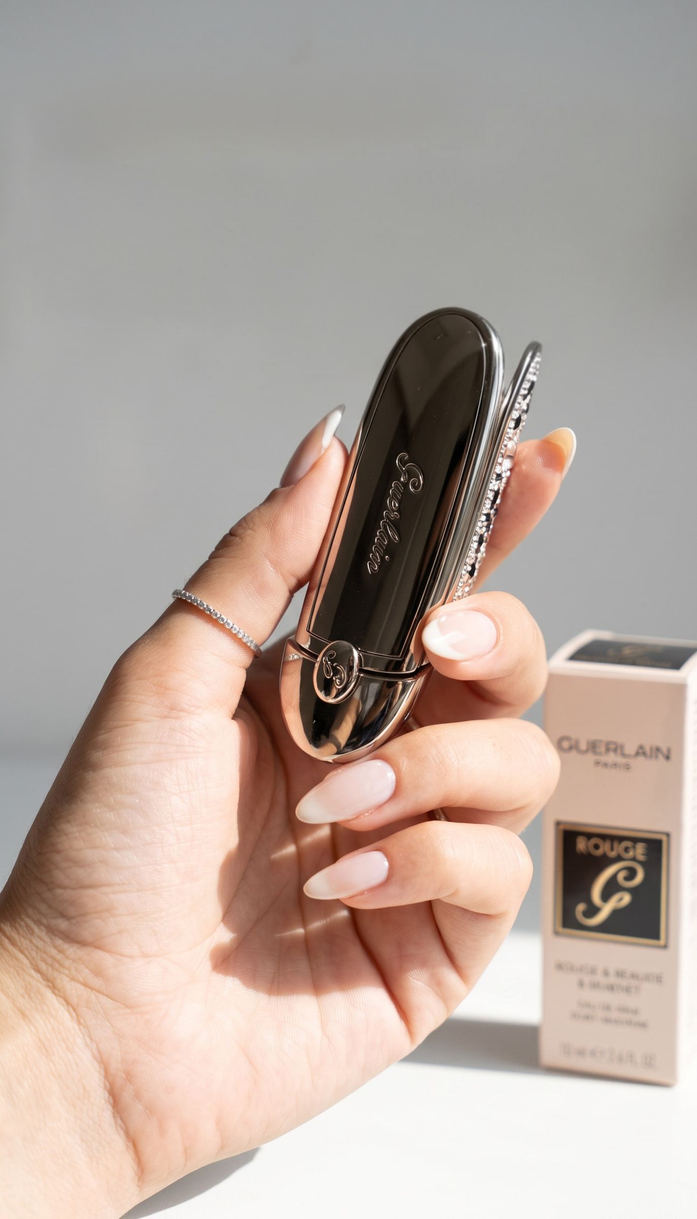Una mano sostiene un estuche de labial Rouge G de Guerlain, con su caja al fondo.