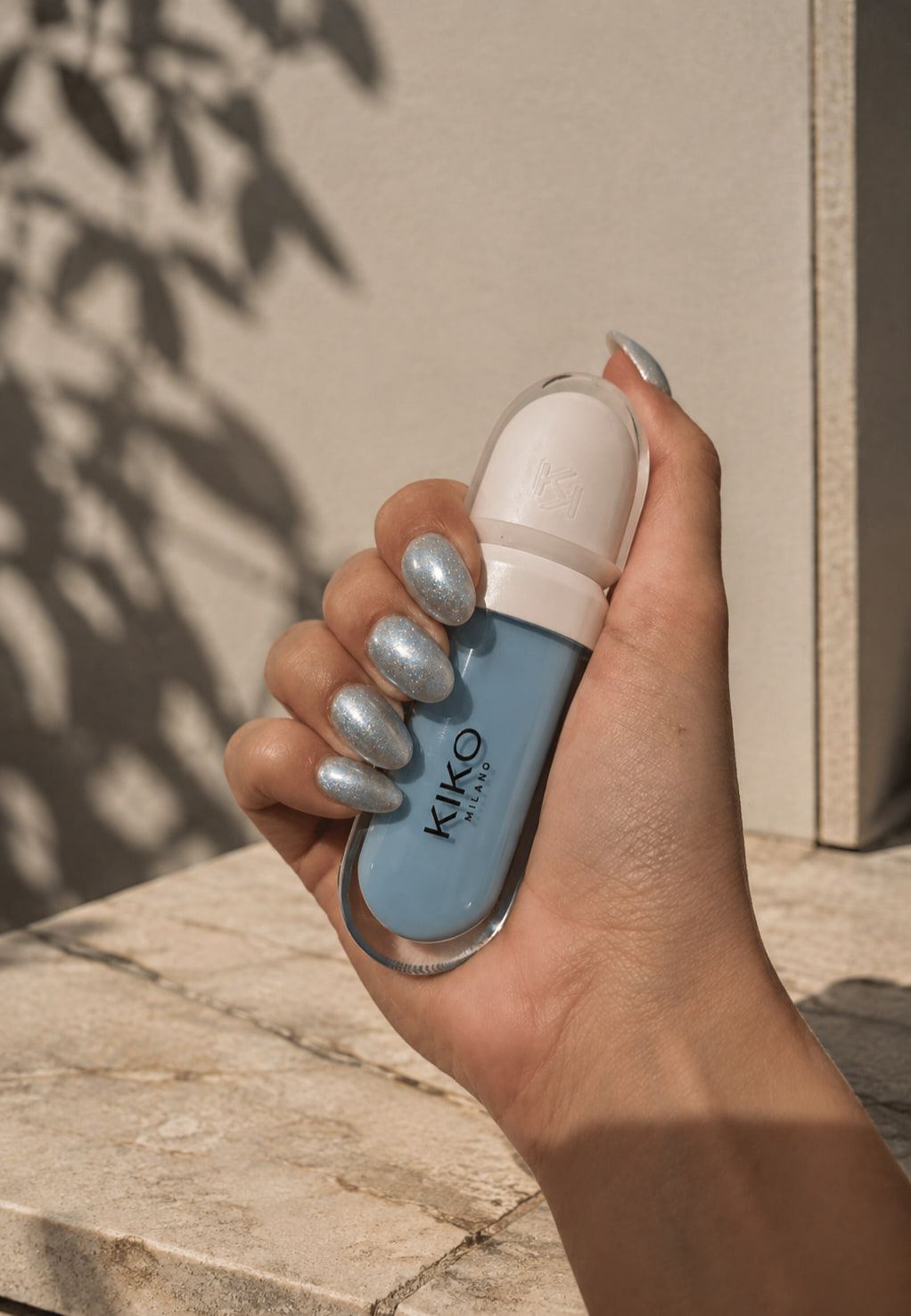 Una mano con uñas metalizadas sostiene un labial azul de Kiko, con sombras de hojas en la pared.