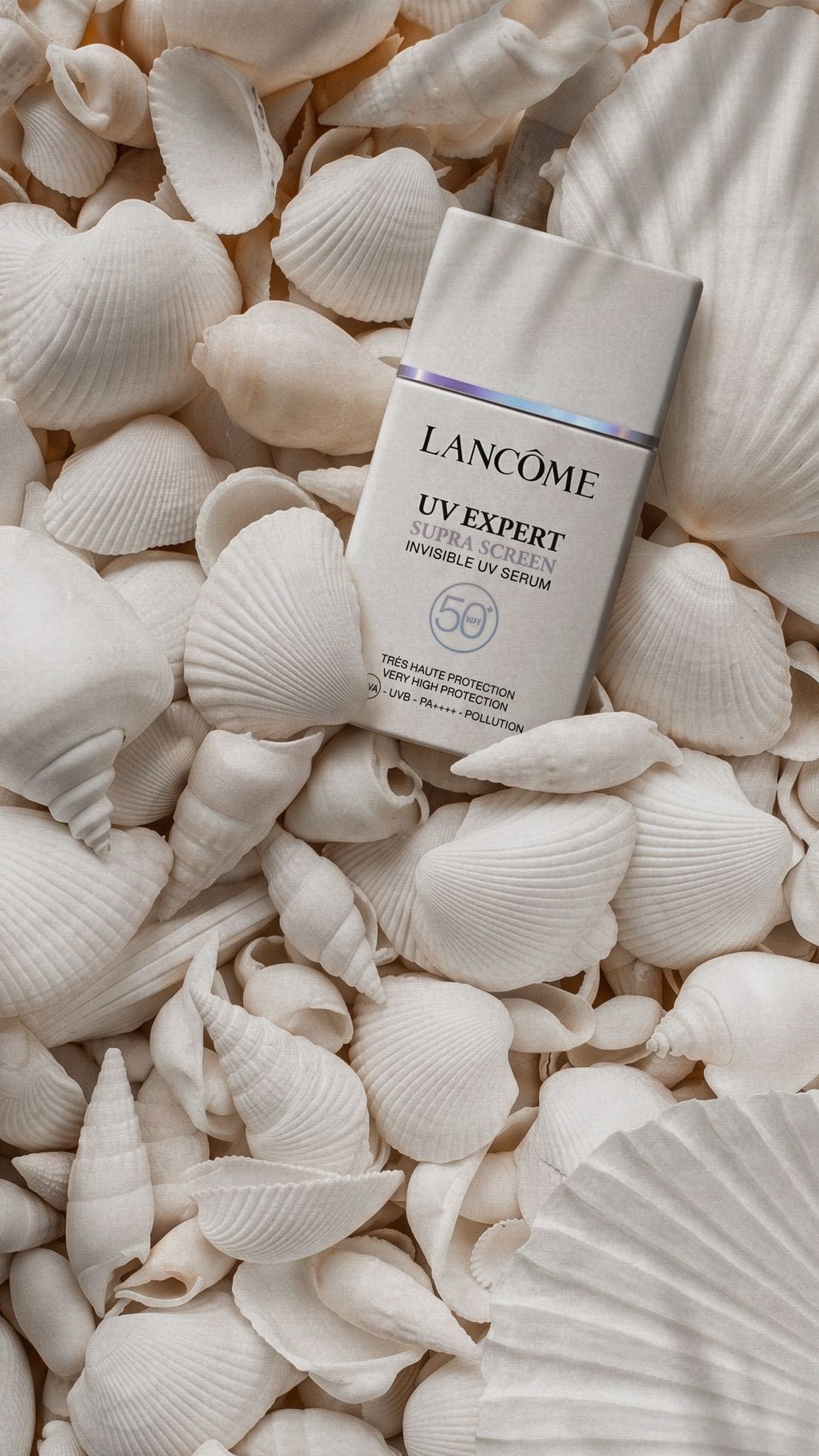 Un sérum solar de Lancôme apoyado sobre un lecho de conchas blancas, con luz suave.