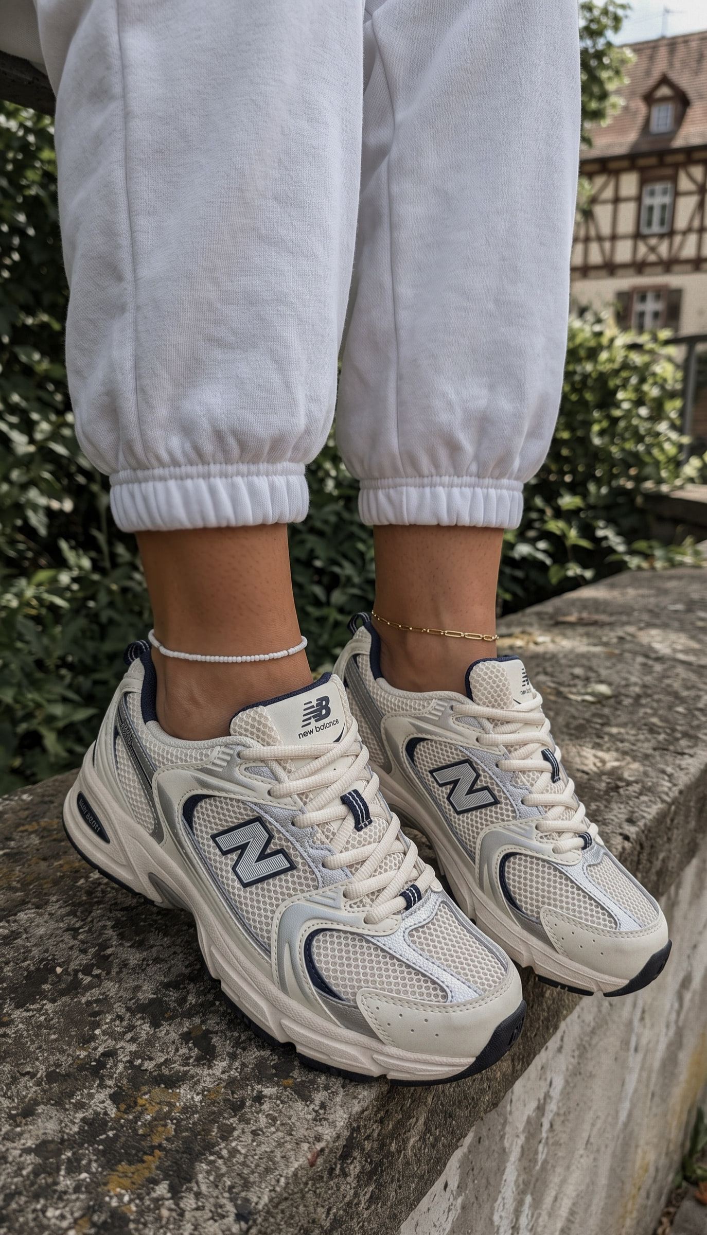 Unas zapatillas New Balance 530 con pantalón de chándal blanco y tobilleras, de pie sobre un muro de piedra.