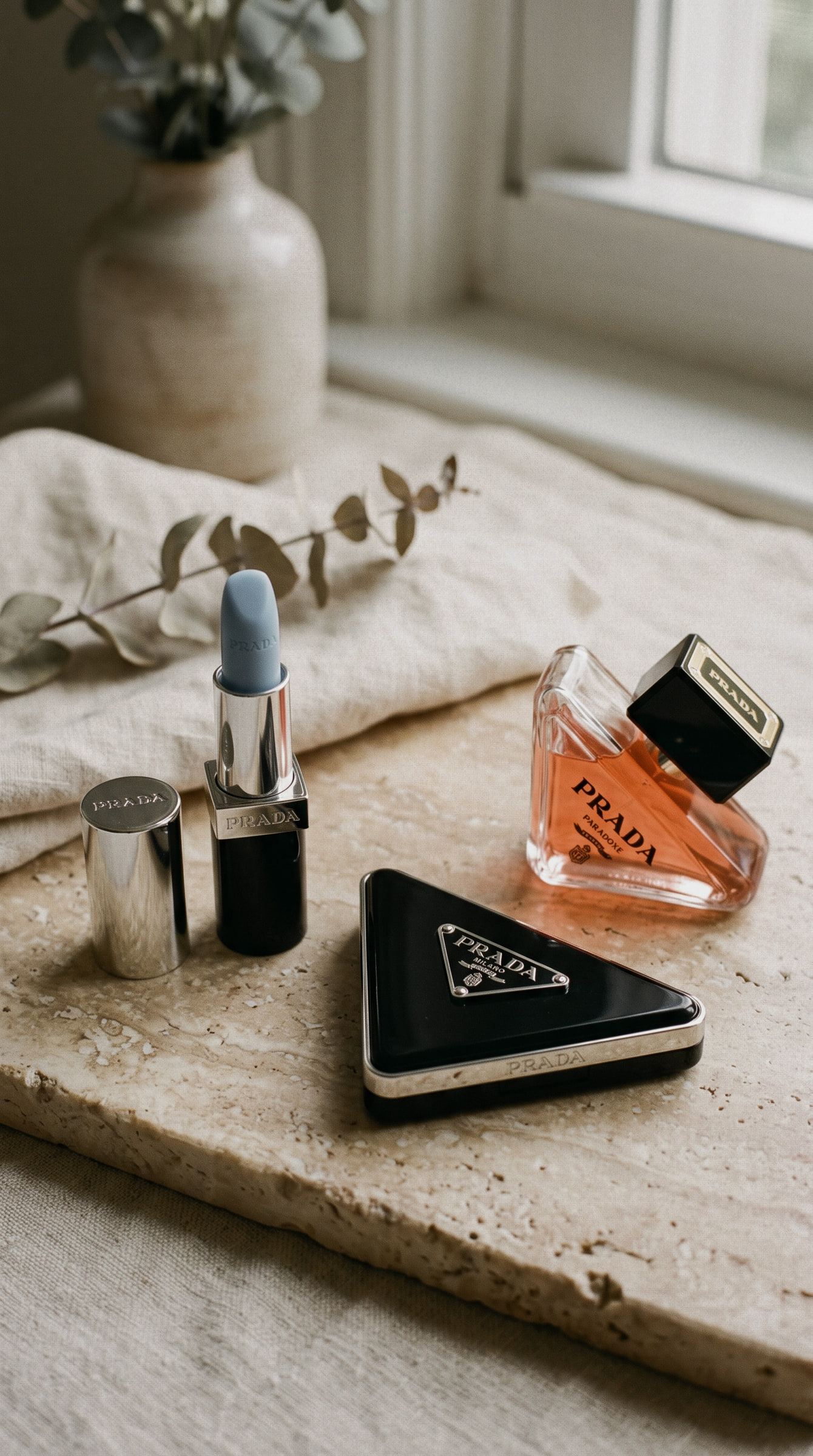 Bodegón de Prada Beauty sobre una tabla de travertino: labial azul, polvos compactos triangulares y perfume.