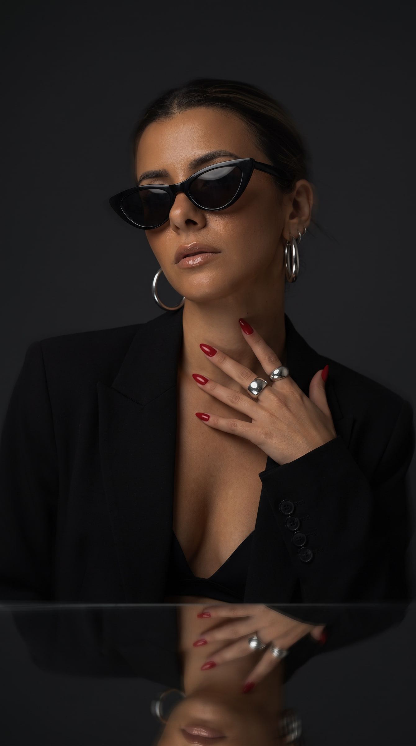 Retrato editorial sobre fondo negro con blazer, gafas de ojo de gato, aros y anillos plateados y uñas rojas reflejadas en una mesa.